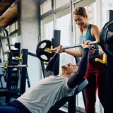 active Therapie und Training Morbach: Ein Mann trainiert an der Langhantel und eine Frau unterstützt ihn dabei.
